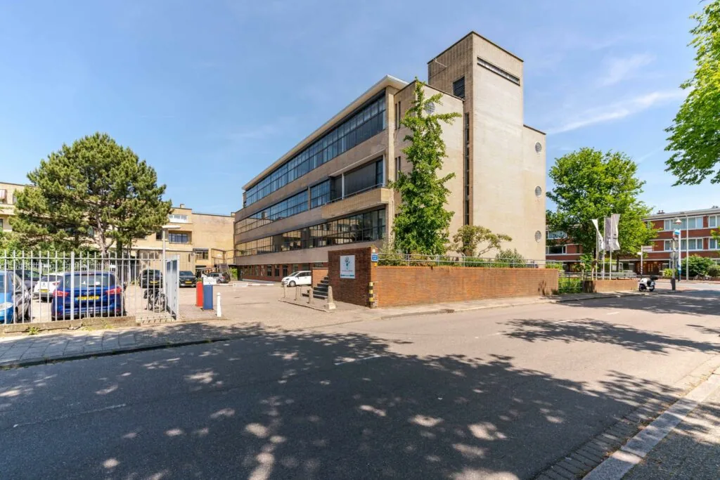 Vooraanzicht van een modernistisch kantoorgebouw aan de Van Alkemadelaan met parkeerplaats en omringende bebouwing.